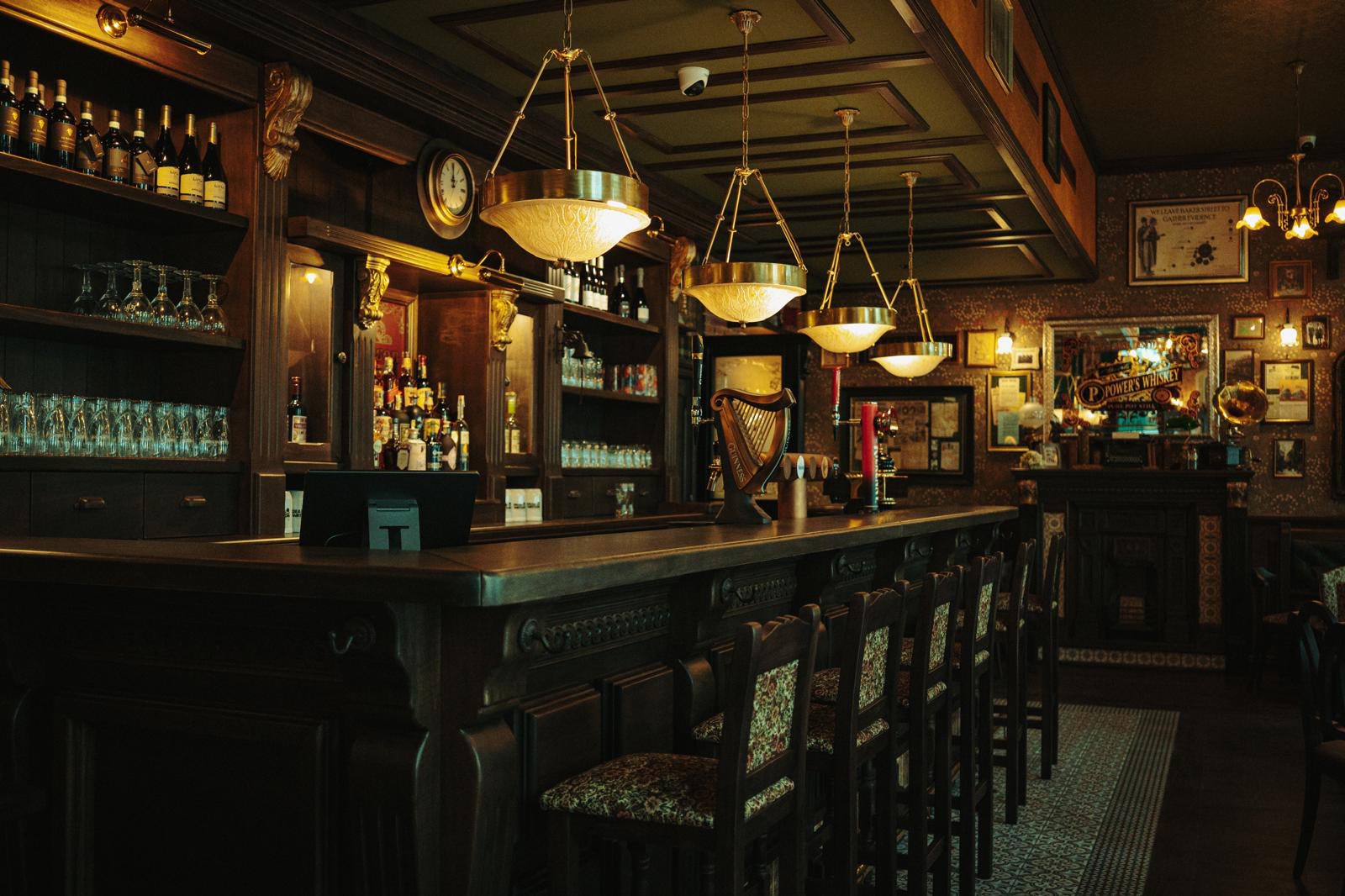 Bar Dear Watson s tmavým dřevem, mosaznými světly, policemi s lahvemi a harfou Guinness.